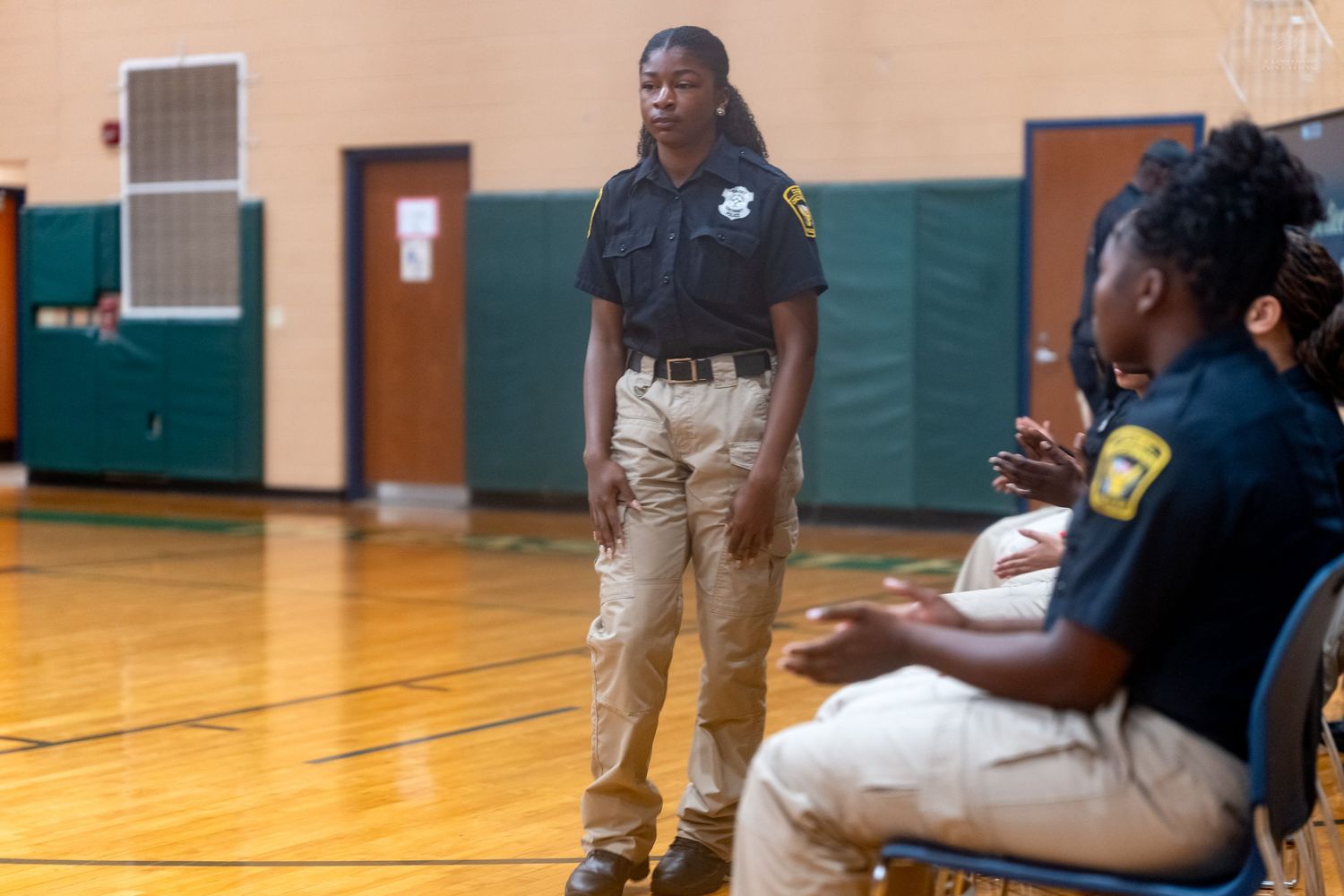 Female police officer stands in gymnasium speaking with seated youth during community engagement session.