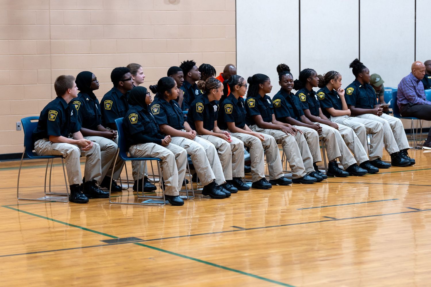 Youth cadets in matching uniforms sit in rows of chairs on a gymnasium floor during a formal ceremony or assembly.
