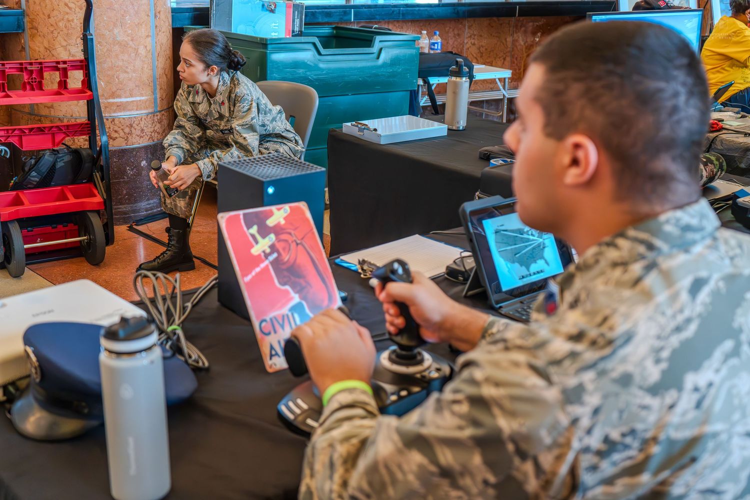 Military personnel in camouflage uniforms operate joystick controls and tablets at an indoor technology demonstration event.