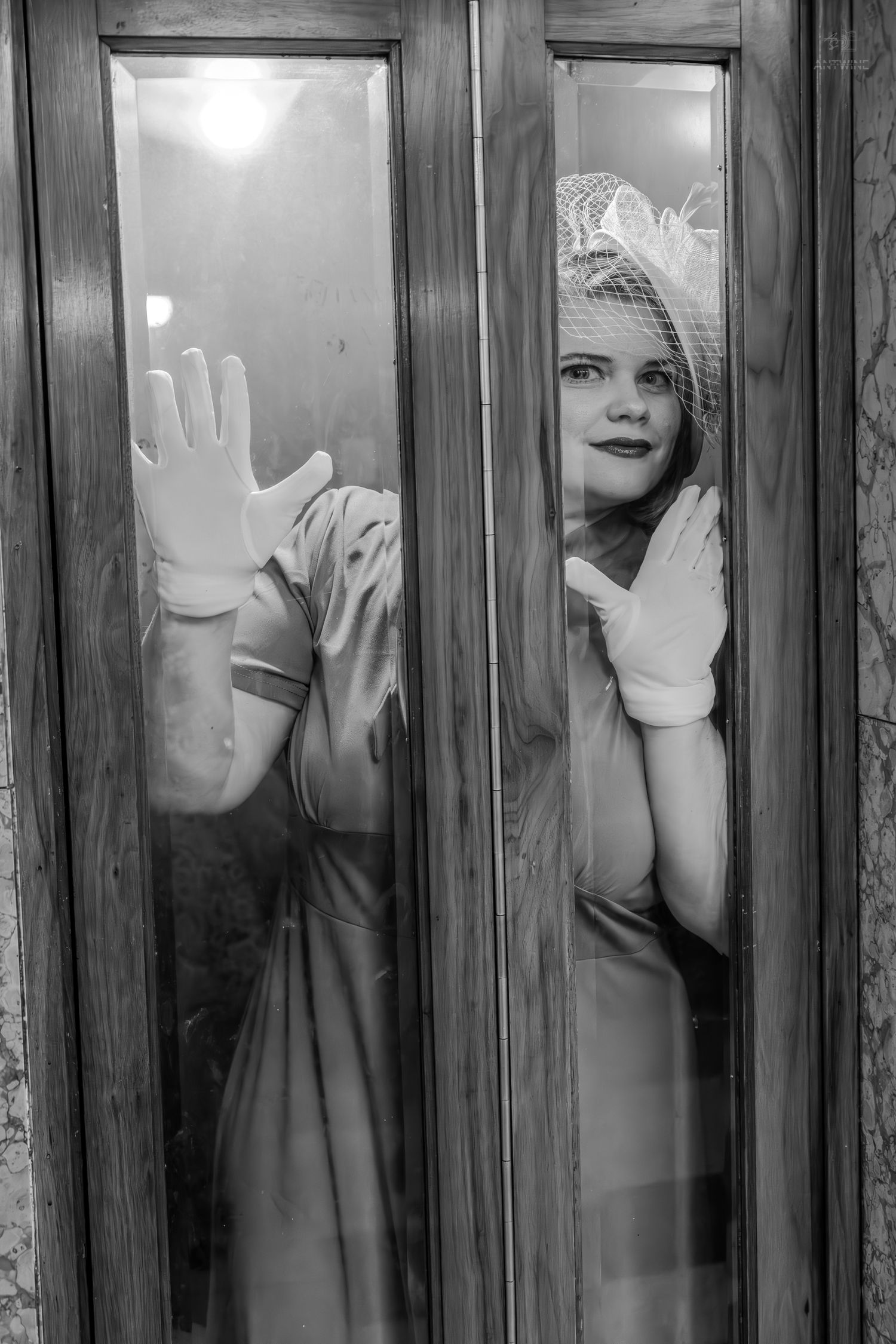 Black and white photo of elegant woman in gloves and veil hat pressing hand against glass door panel.