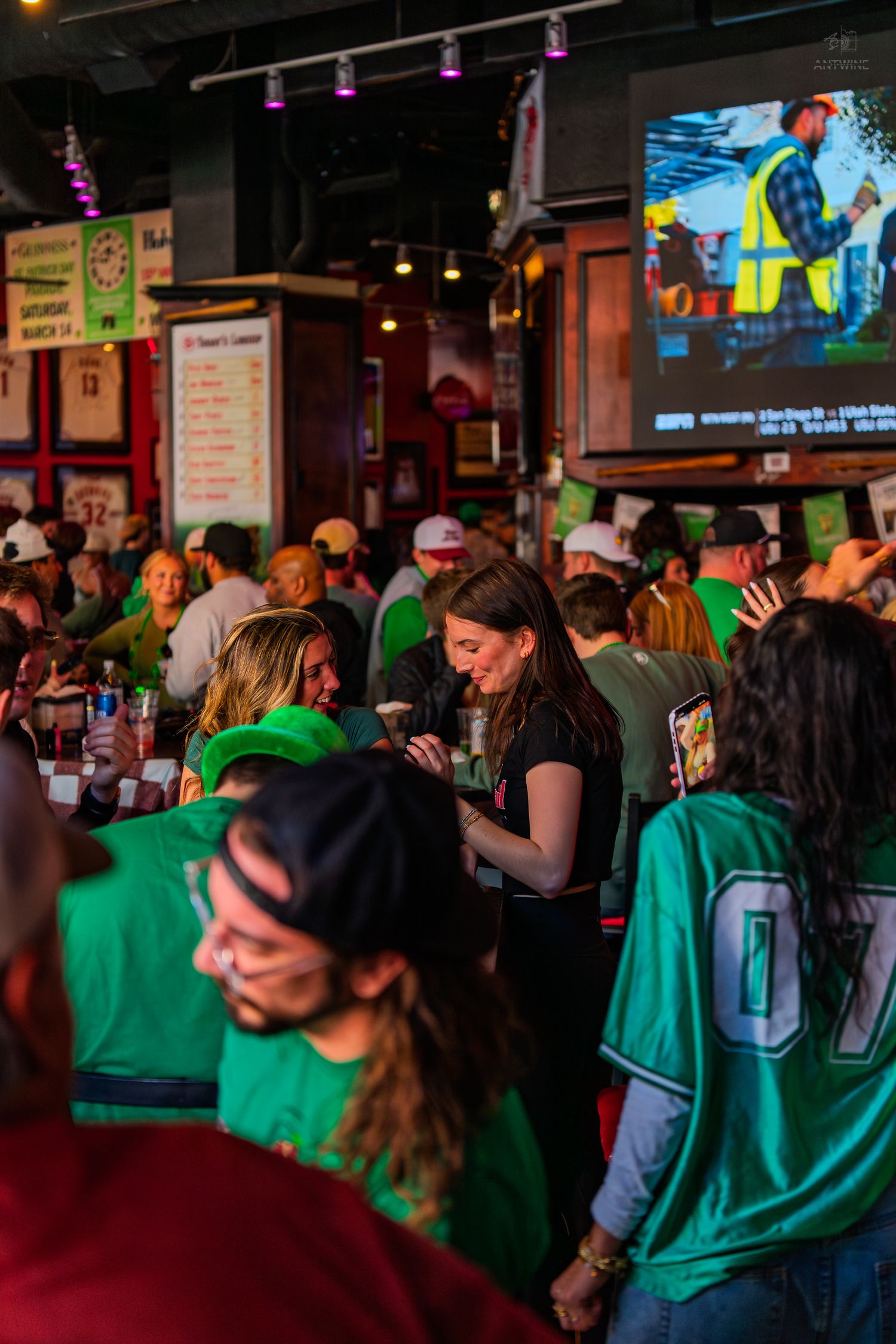 Busy sports bar packed with patrons wearing green jerseys celebrating St. Patrick's Day while a server takes orders.