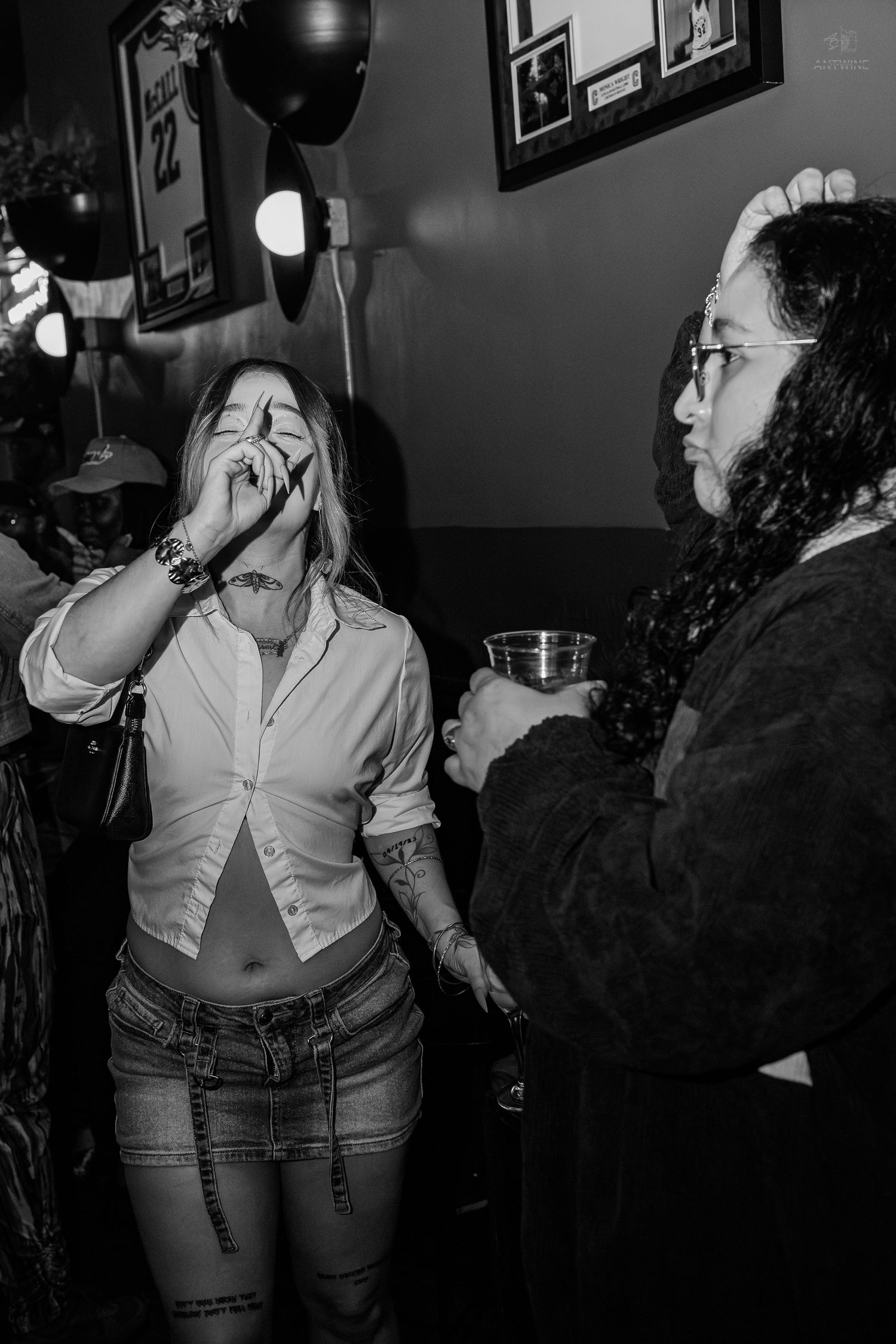 Black and white photo of two people socializing at a bar, woman holding drink to face, tattooed man holding cup.