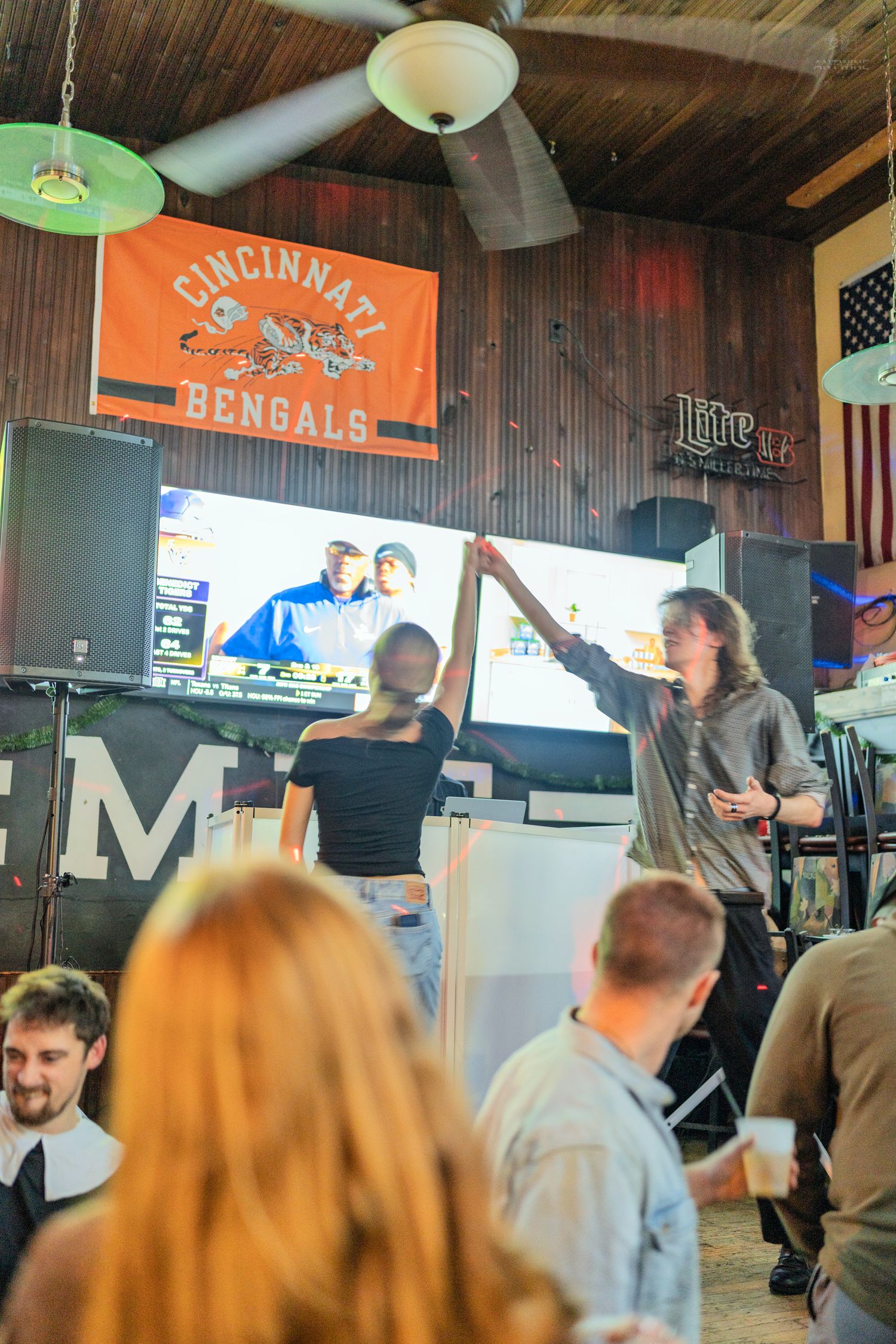 People dancing inside a bar featuring a Cincinnati Bengals banner and large TV screen.