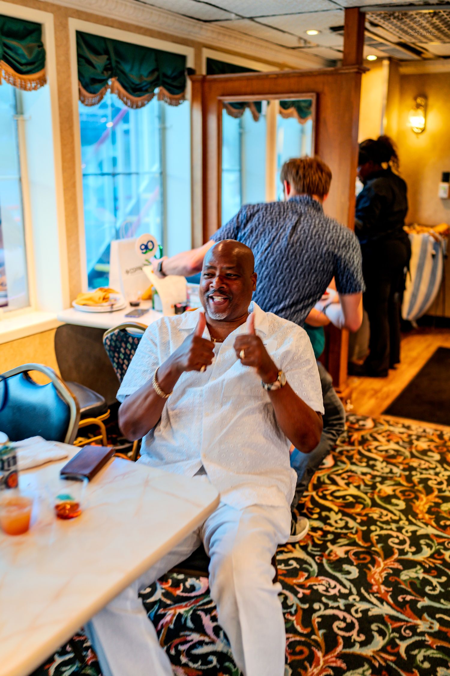 Man in white outfit giving thumbs up while seated at table in ornate dining room.