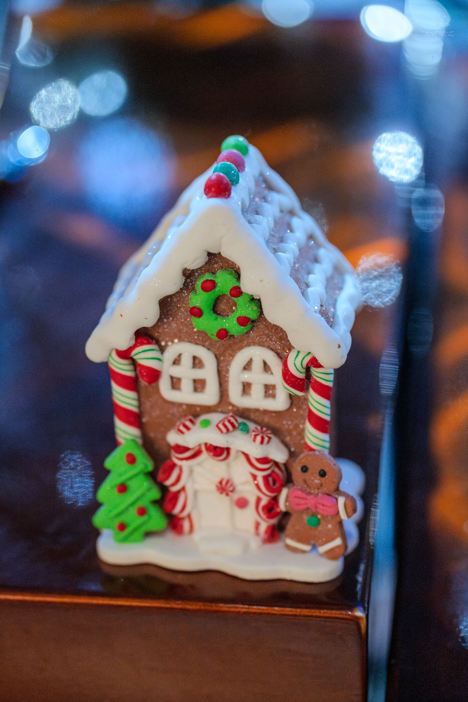 A decorative gingerbread house with white icing, candy canes, and a gingerbread man figurine against a bokeh light background.