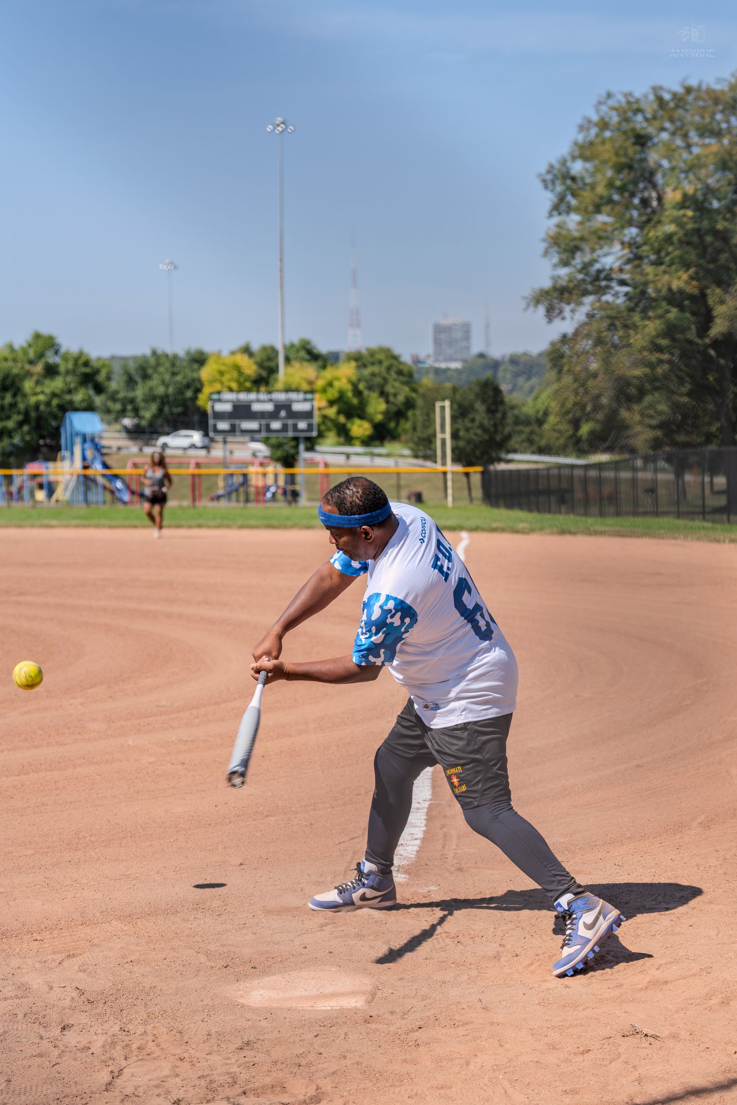 A softball batter swings at a yellow ball during an outdoor game on a dirt field on a sunny day.