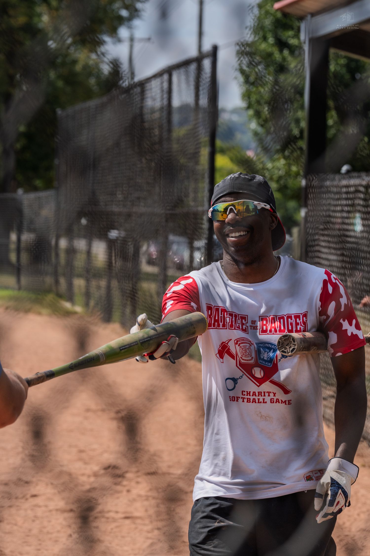 Smiling baseball player holding a bat at a charity softball game wearing a red camo jersey and sunglasses.