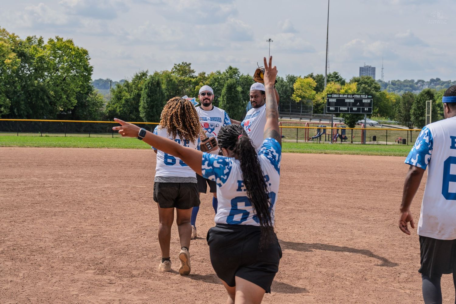 Softball players in blue and white jerseys celebrate on a dirt field, with one player flashing a peace sign.