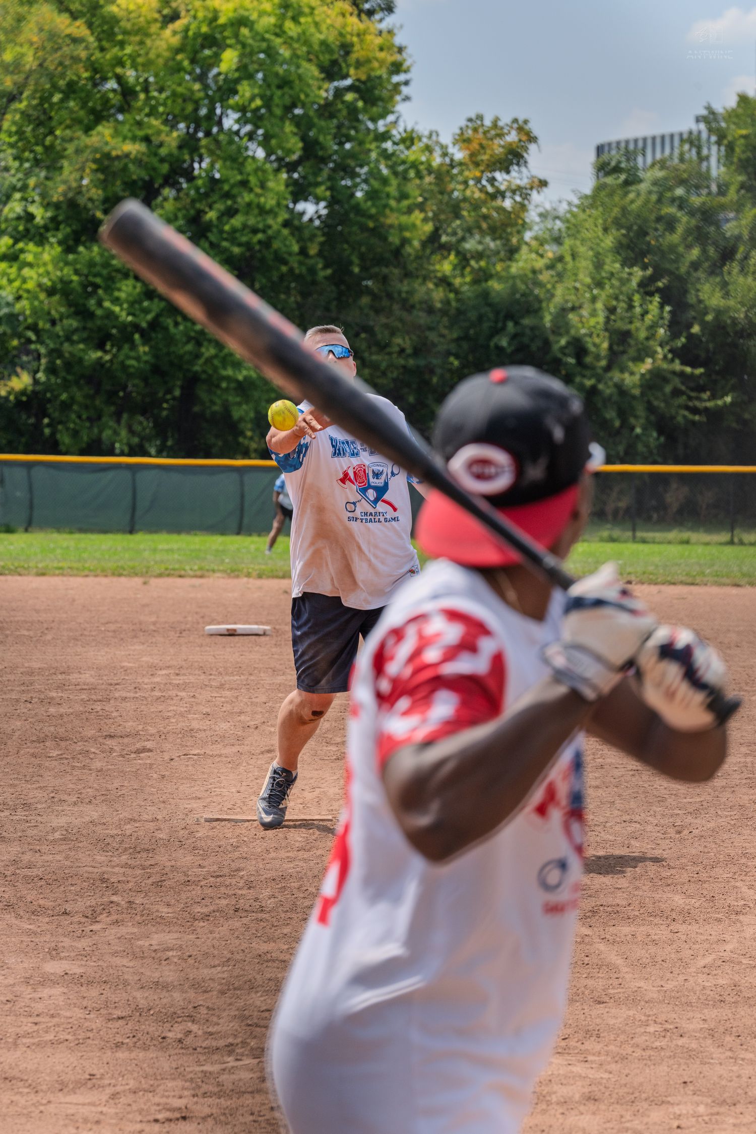 A pitcher releases the ball as a batter in a red jersey prepares to swing in a softball game.