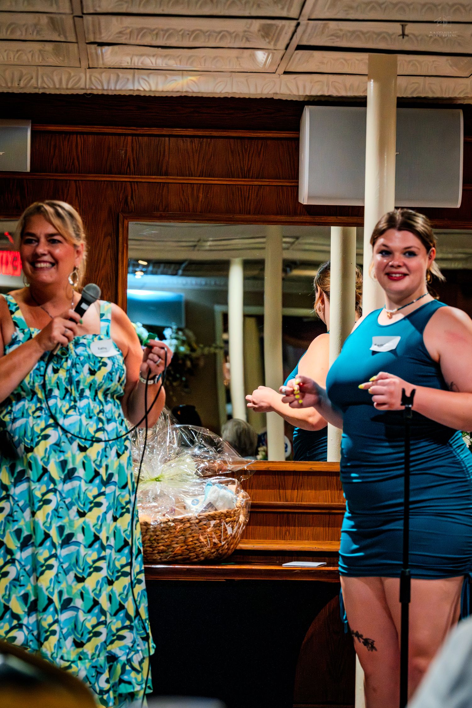 Two women in blue dresses smile on stage, one holding a microphone at an indoor event.