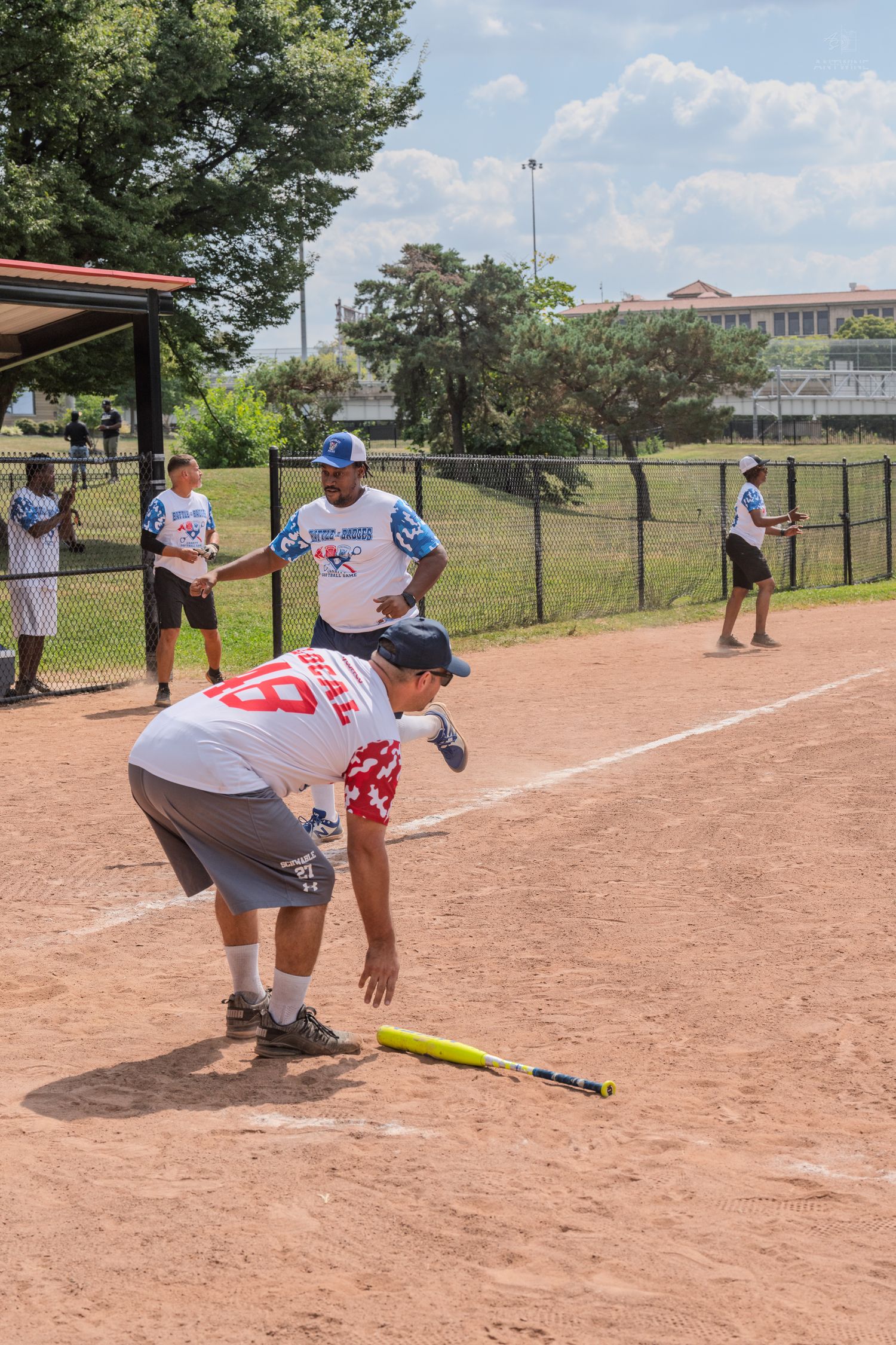 A softball player bends to pick up a yellow bat on a dirt field during a casual game.