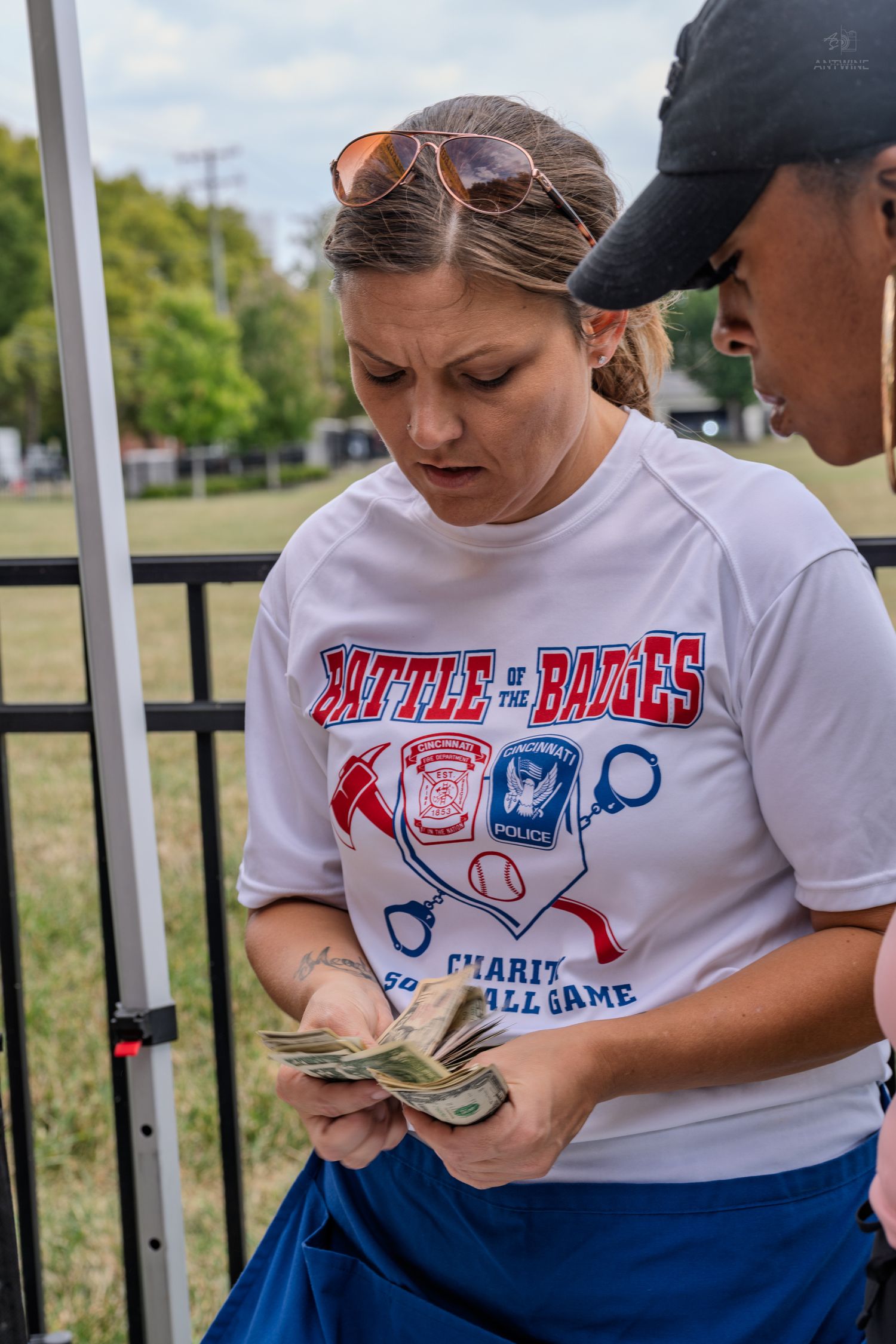 A woman in a Battle of the Badges charity softball shirt counts cash money under a tent at an outdoor event.