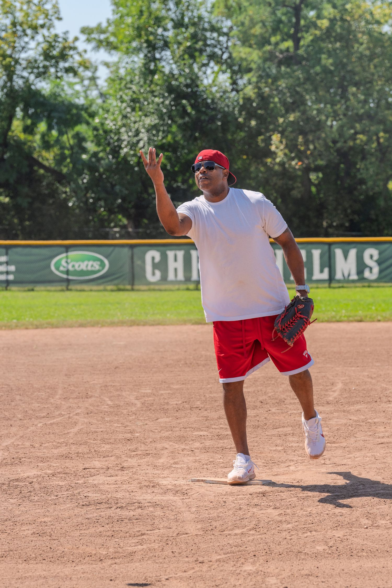 Man in red shorts and white shirt throwing a softball on a baseball diamond on a sunny day.