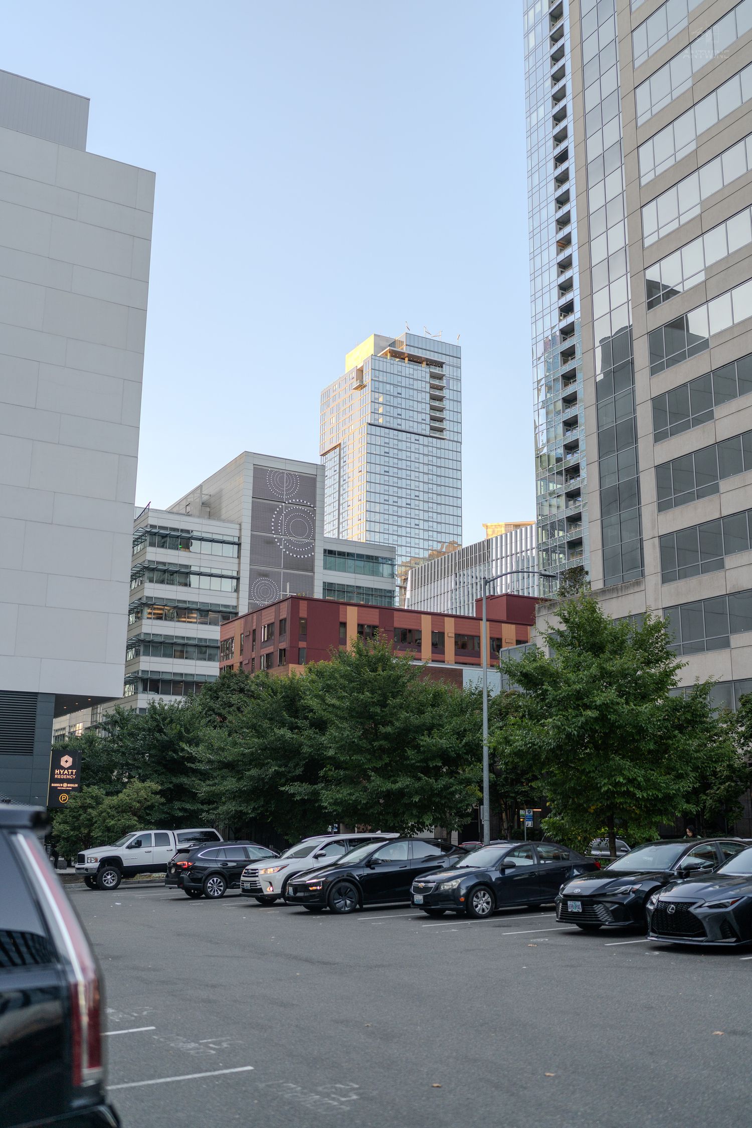 Urban parking lot surrounded by modern skyscrapers and green trees under a clear blue sky.