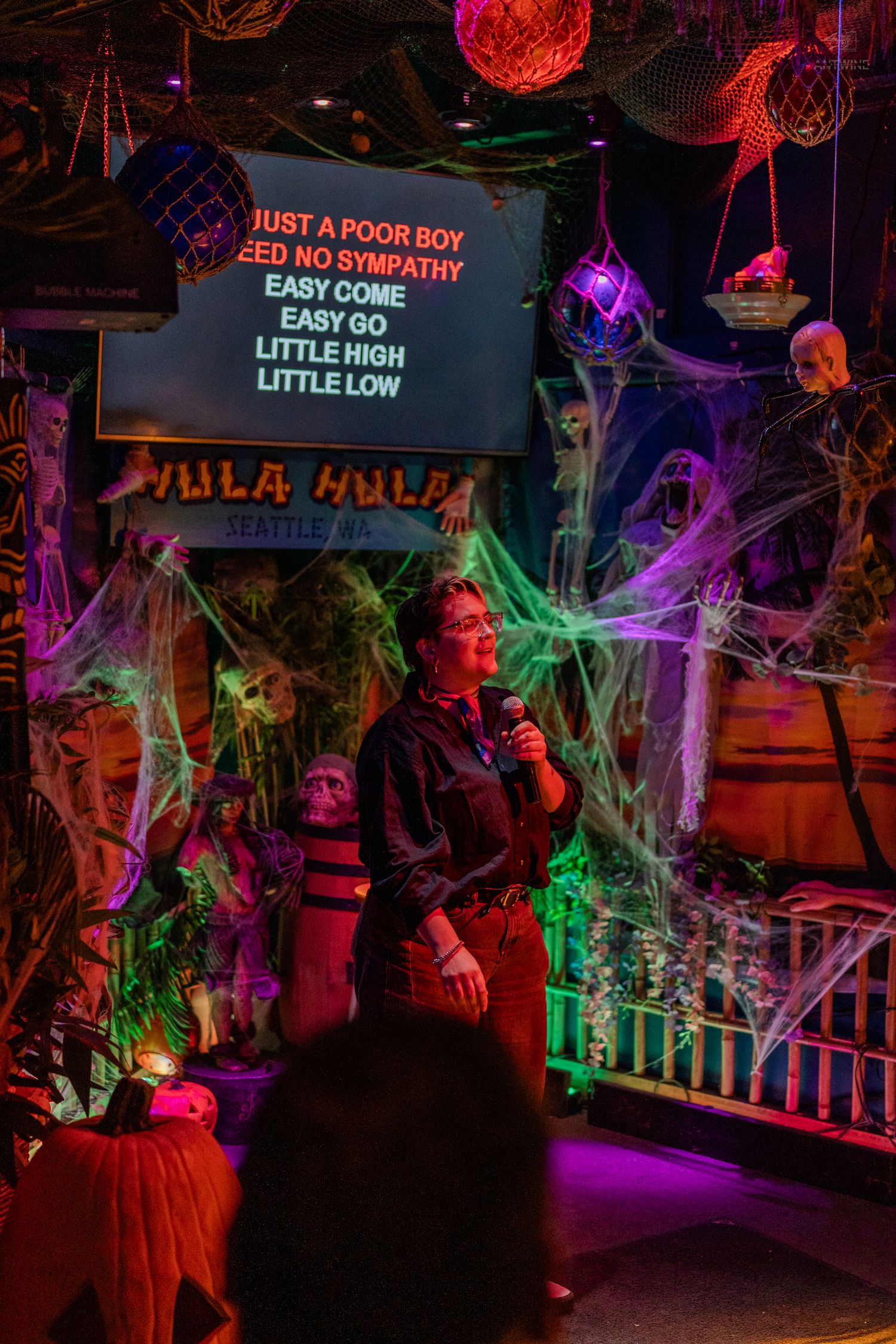 Person singing karaoke at Hula Hula bar in Seattle with Halloween decorations and Bohemian Rhapsody lyrics on screen.