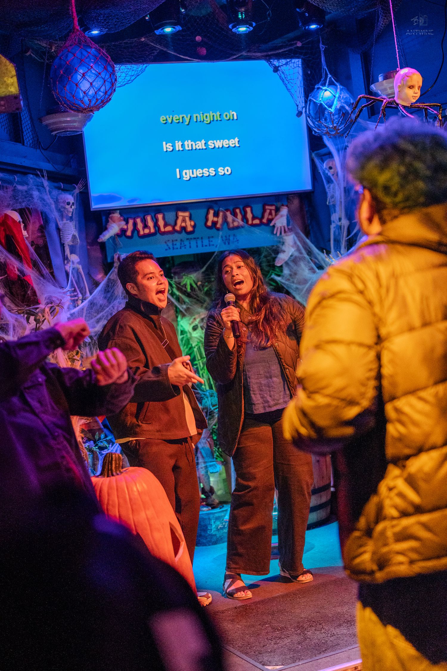 Two people singing karaoke on a Halloween-decorated stage at Hula Hula bar in Seattle, WA.