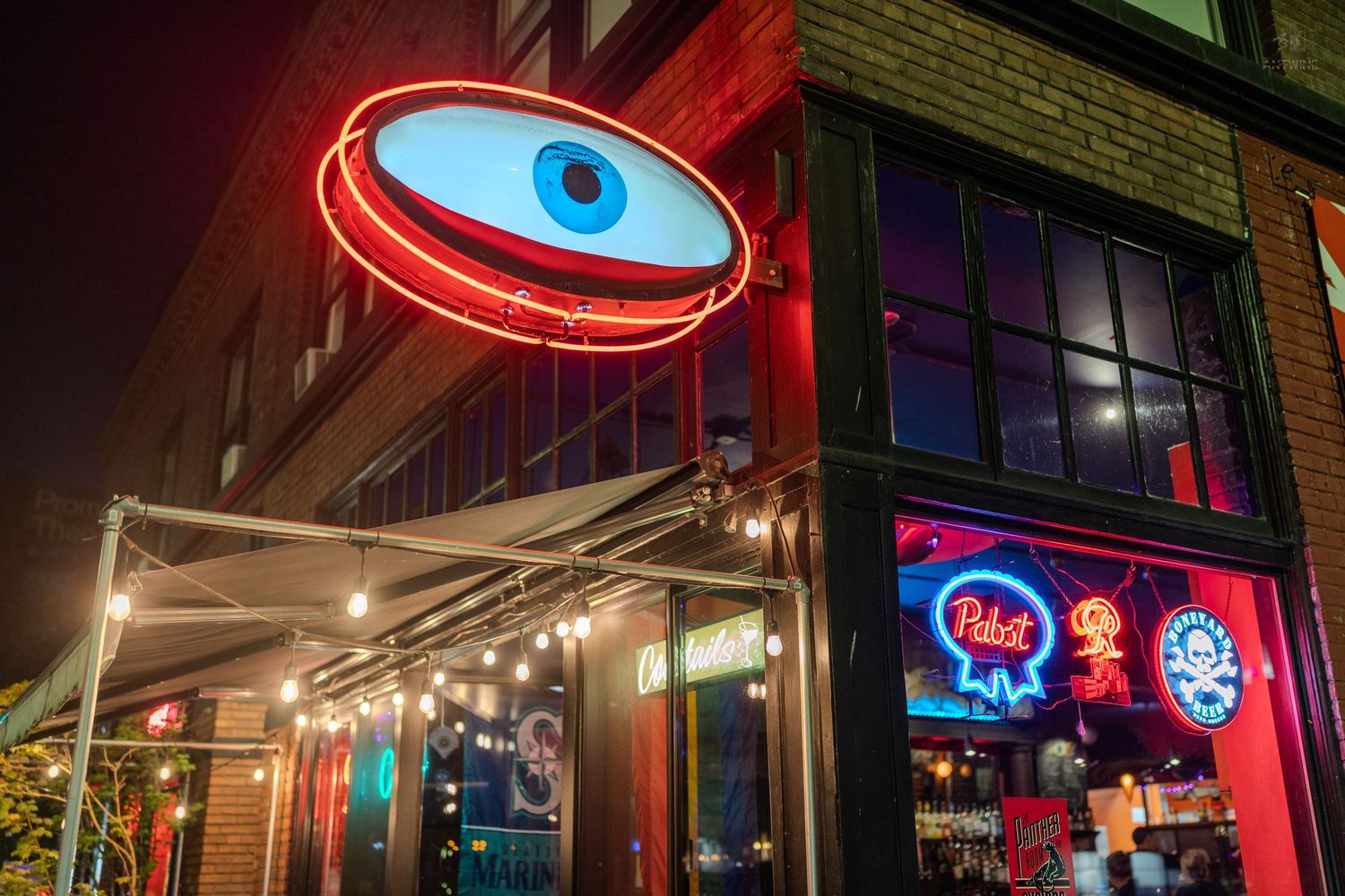 Neon eye sign glows above a bar entrance at night with Pabst Blue Ribbon signs illuminating the storefront.