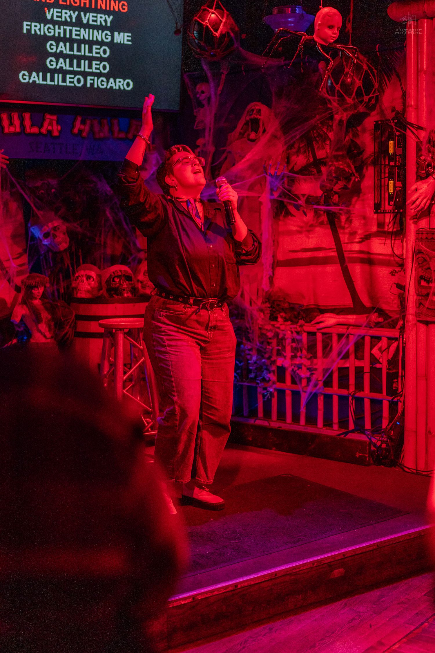 Person singing Bohemian Rhapsody karaoke on a red-lit Halloween-decorated stage with spiderwebs.
