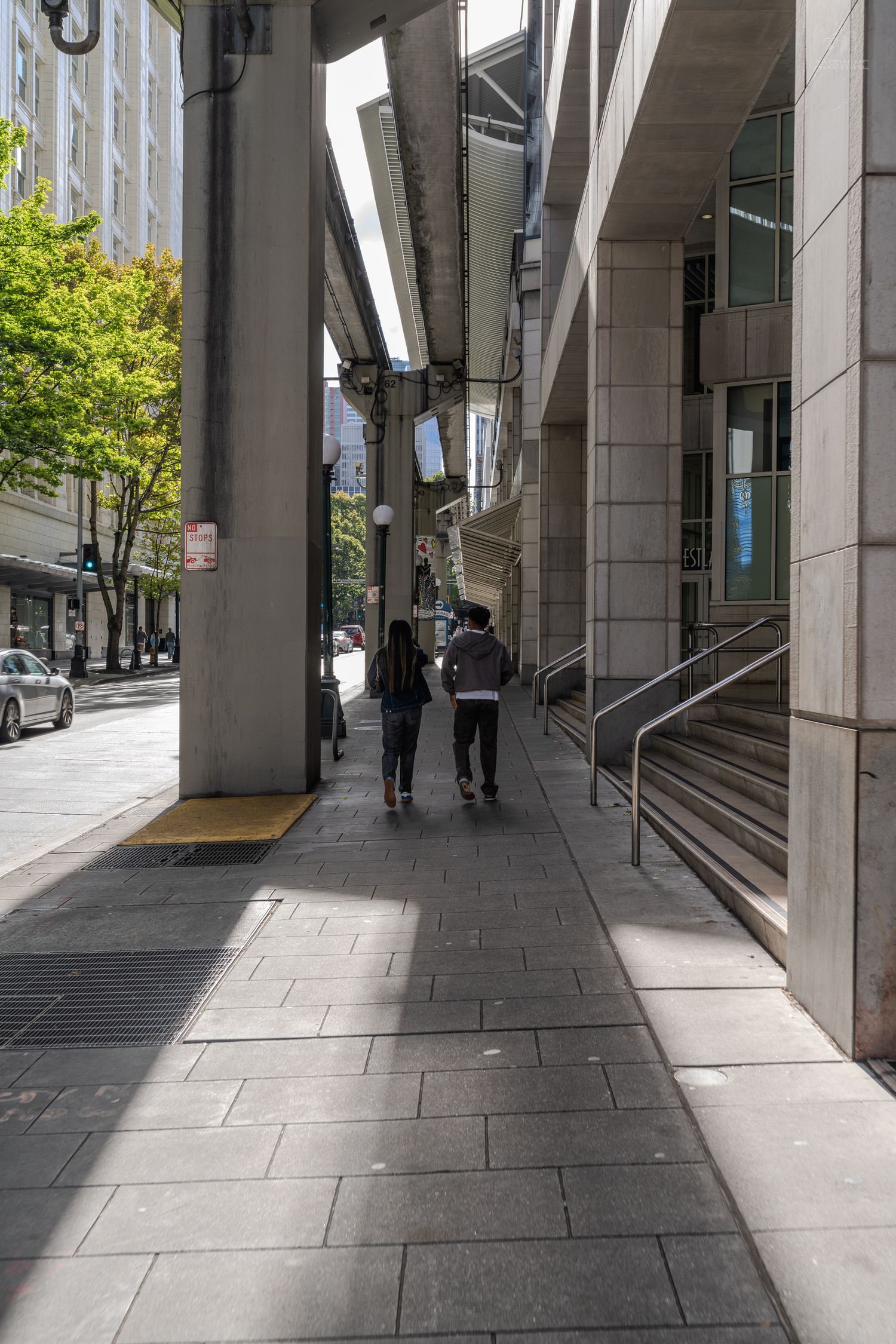 Two pedestrians walking along shaded urban sidewalk beneath elevated structure with concrete pillars in downtown Seattle.