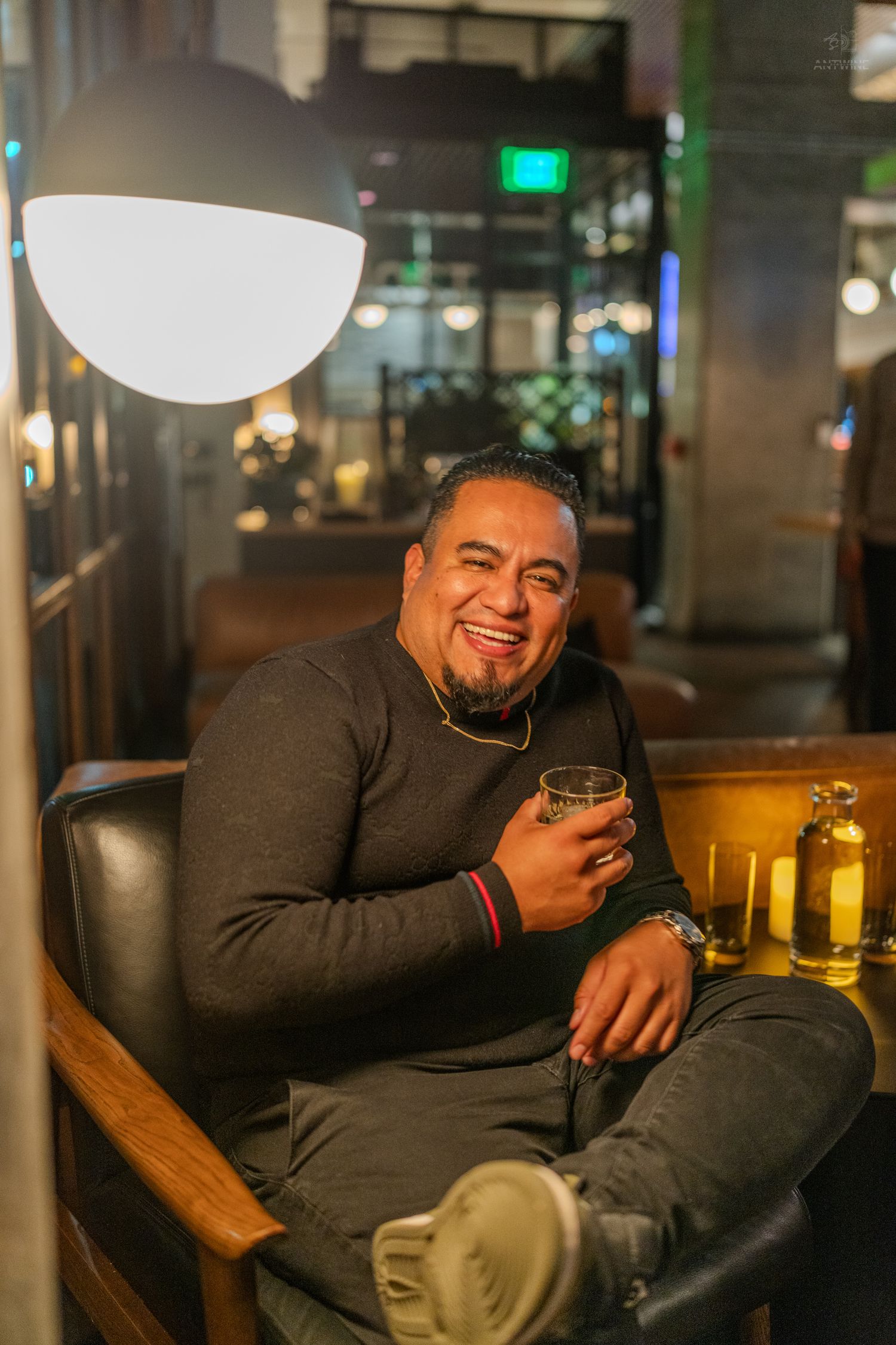 A cheerful man in a black sweater smiles while holding a drink in a stylish lounge setting.