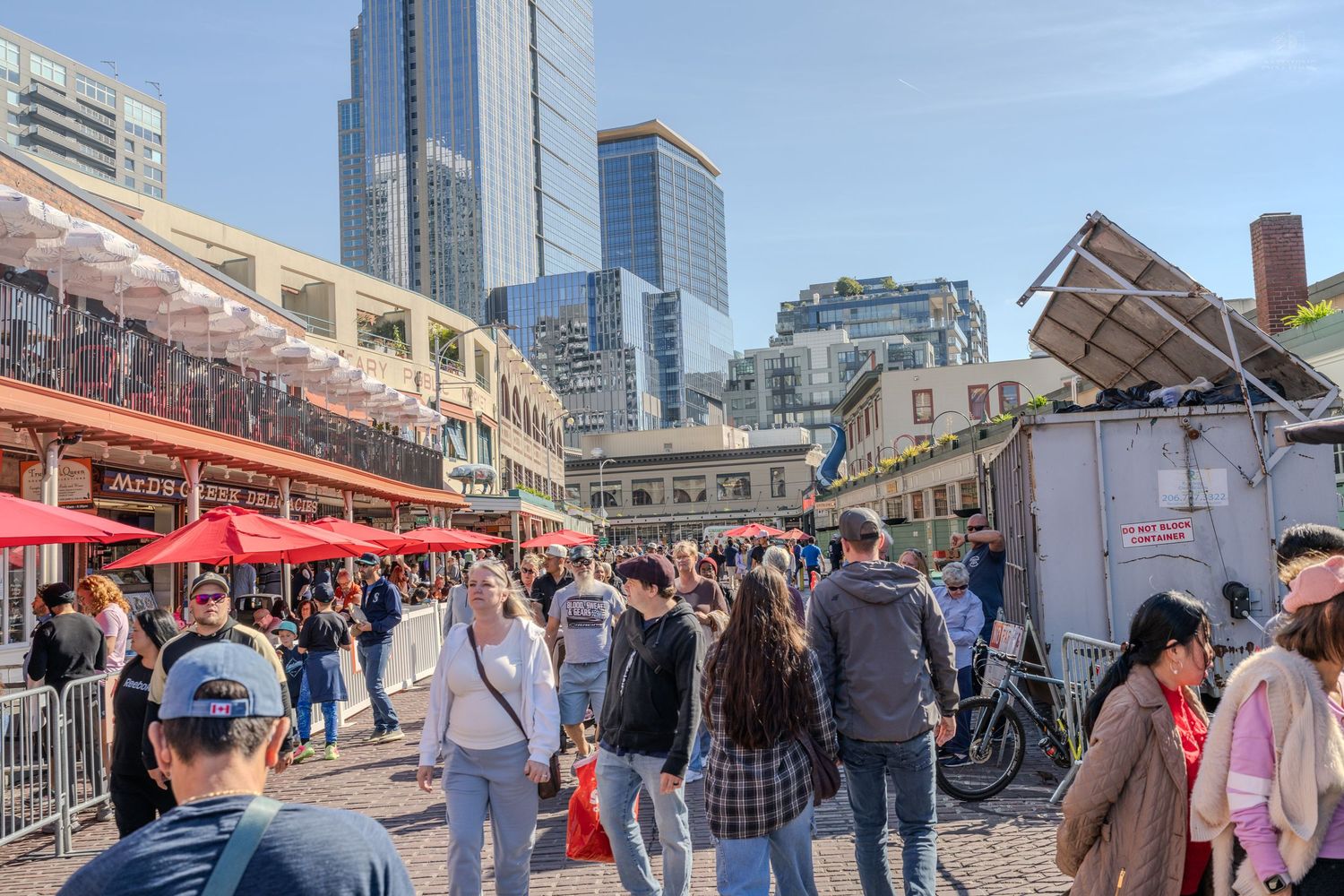 Crowded outdoor market near Seattle skyscrapers with vendors, visitors, and red umbrellas on a sunny day.