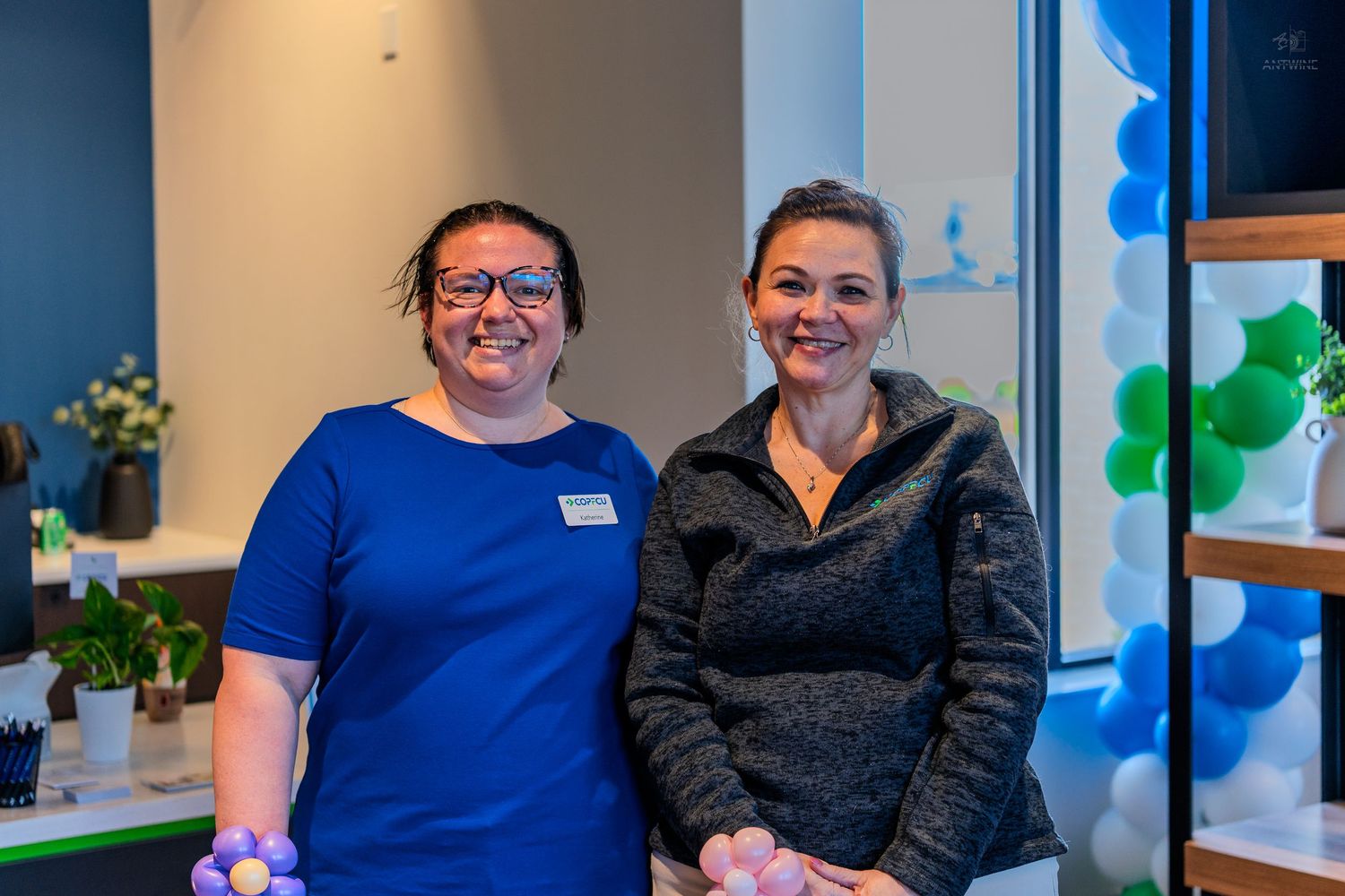 Two smiling women pose together in a bright modern office decorated with colorful balloons and plants.