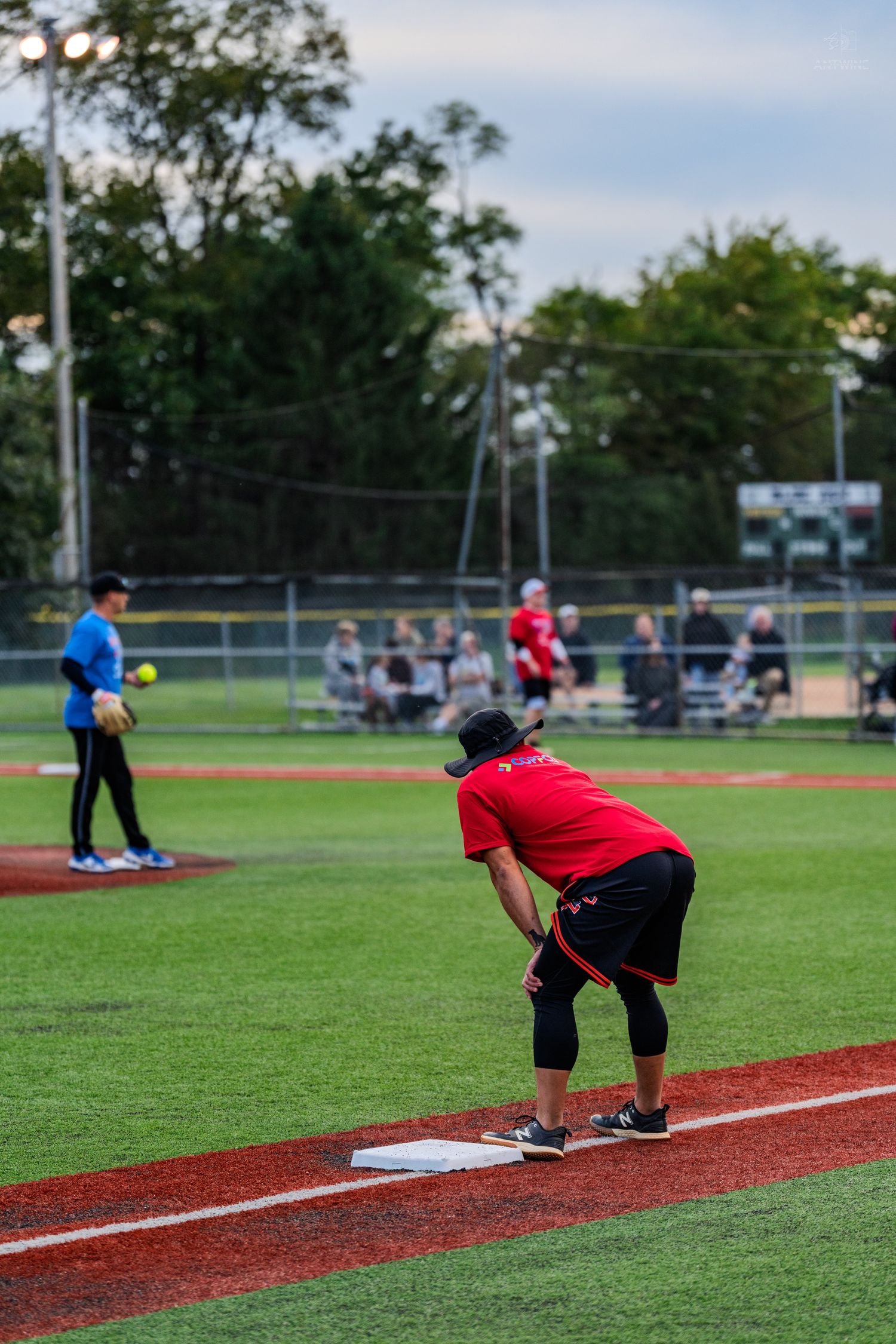 Player in red uniform crouches at third base during an evening recreational softball game on a well-lit field.