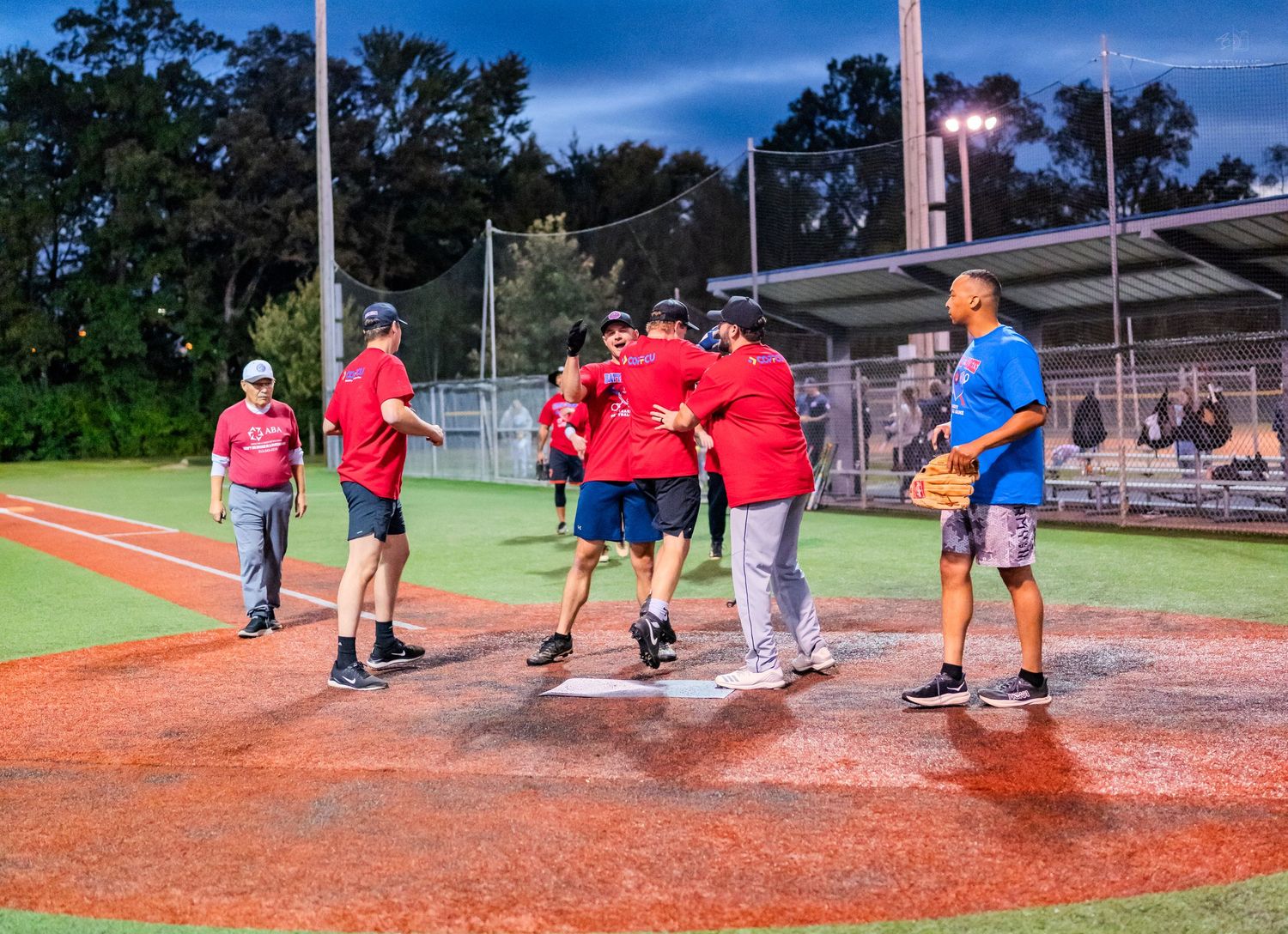 Group of people in red shirts celebrating on a baseball diamond at night under field lights.
