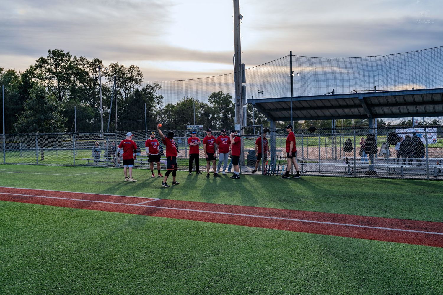 Youth football team in red uniforms gathered on a green field at dusk near bleachers and chain-link fence.