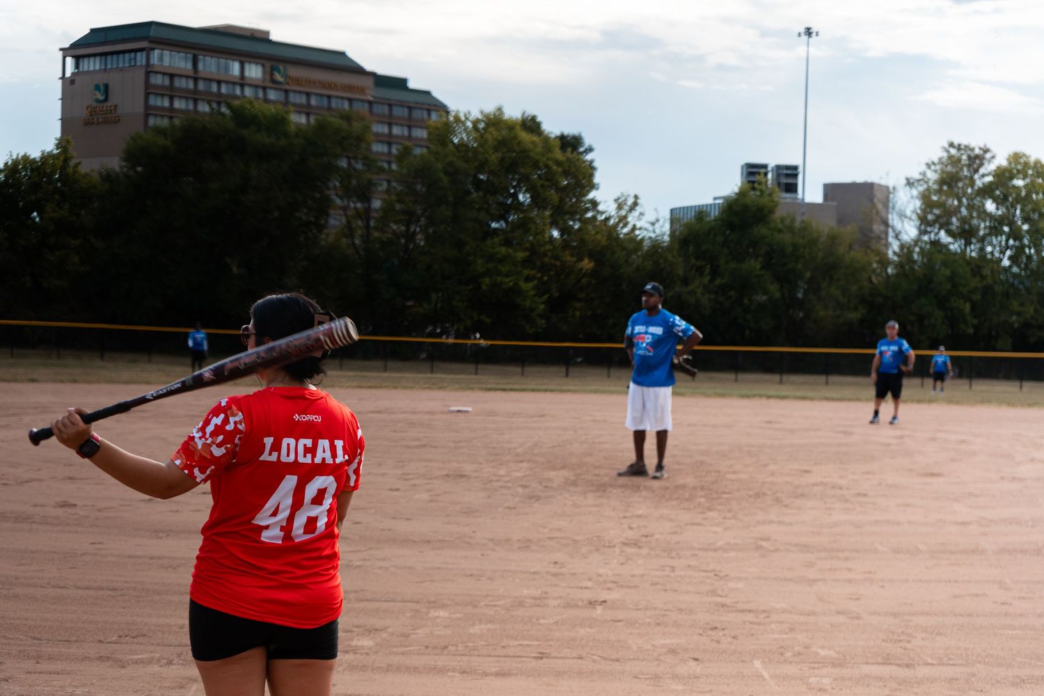 A batter in a red Local 48 jersey prepares to hit during an outdoor softball game on a dirt field.