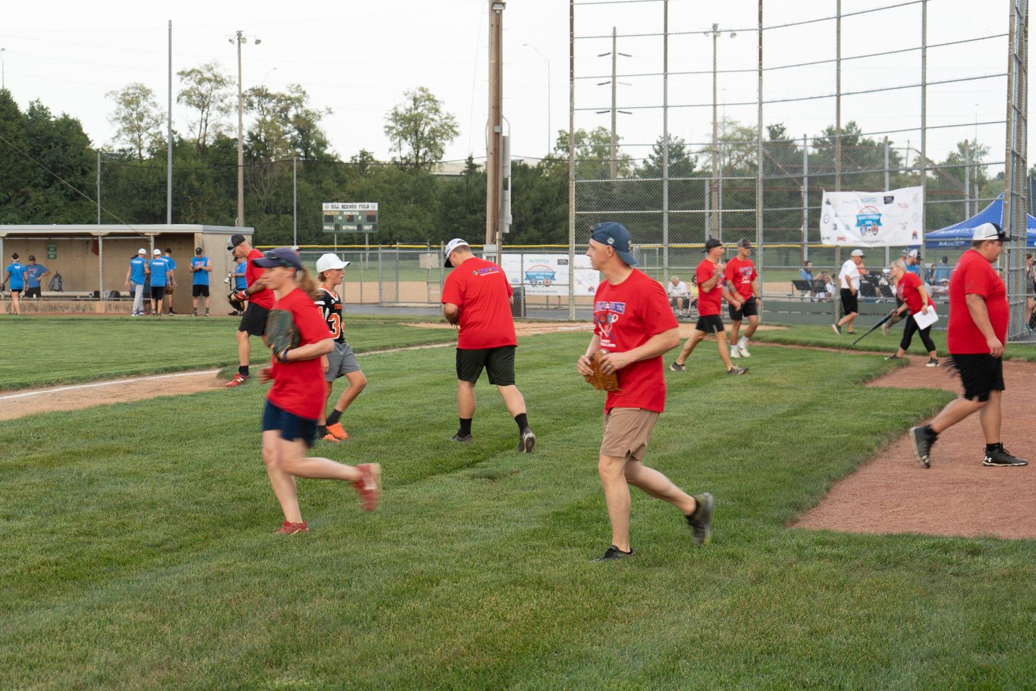 Players in red shirts run the bases on a green baseball field during an outdoor recreational game event.