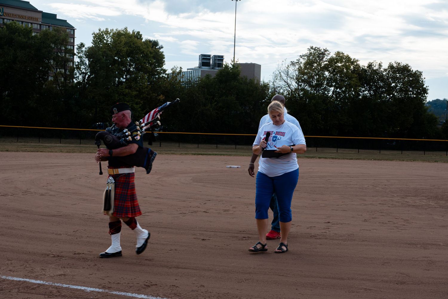 A bagpiper in traditional Scottish attire performs on a baseball field alongside a woman holding a microphone outdoors.