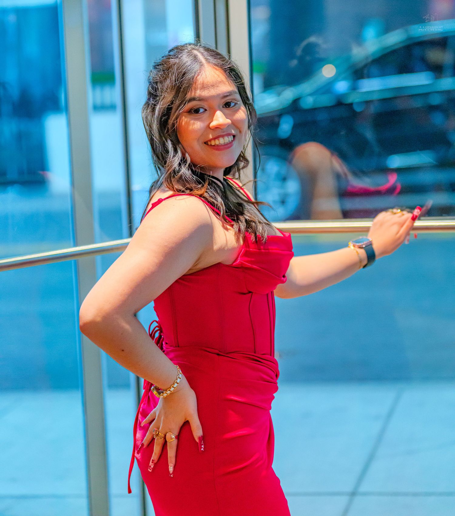Young woman in a red dress posing gracefully by a metal railing, smiling outdoors in an urban setting.