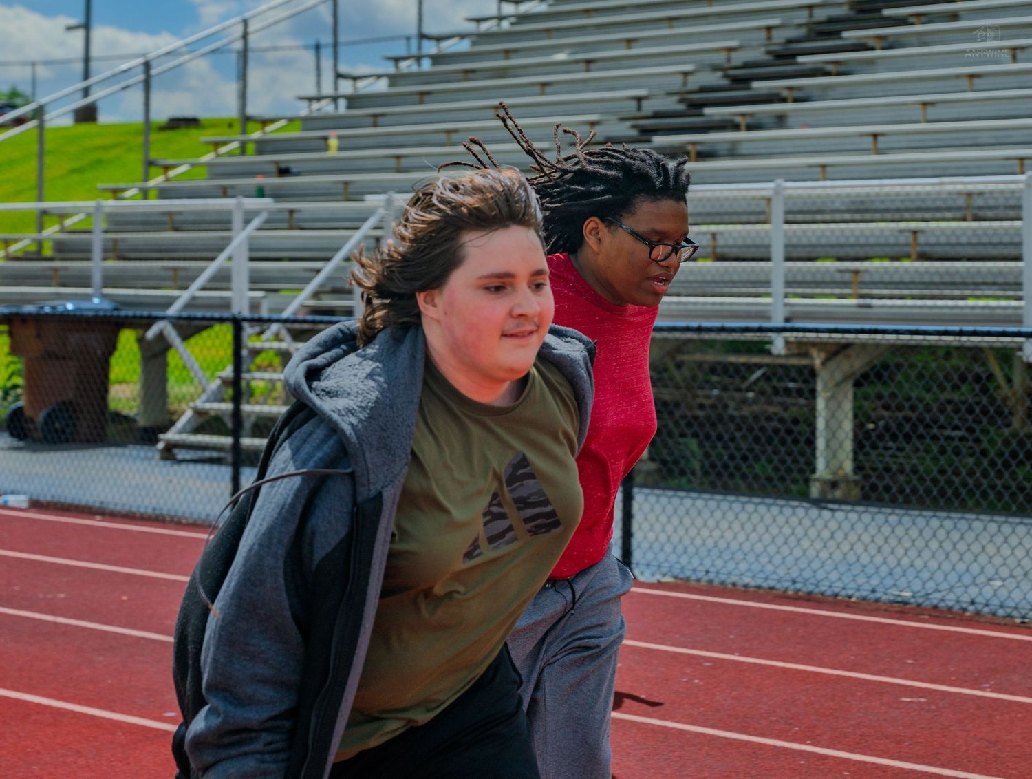 Two students running on a red track with bleachers in the background during outdoor physical education class.