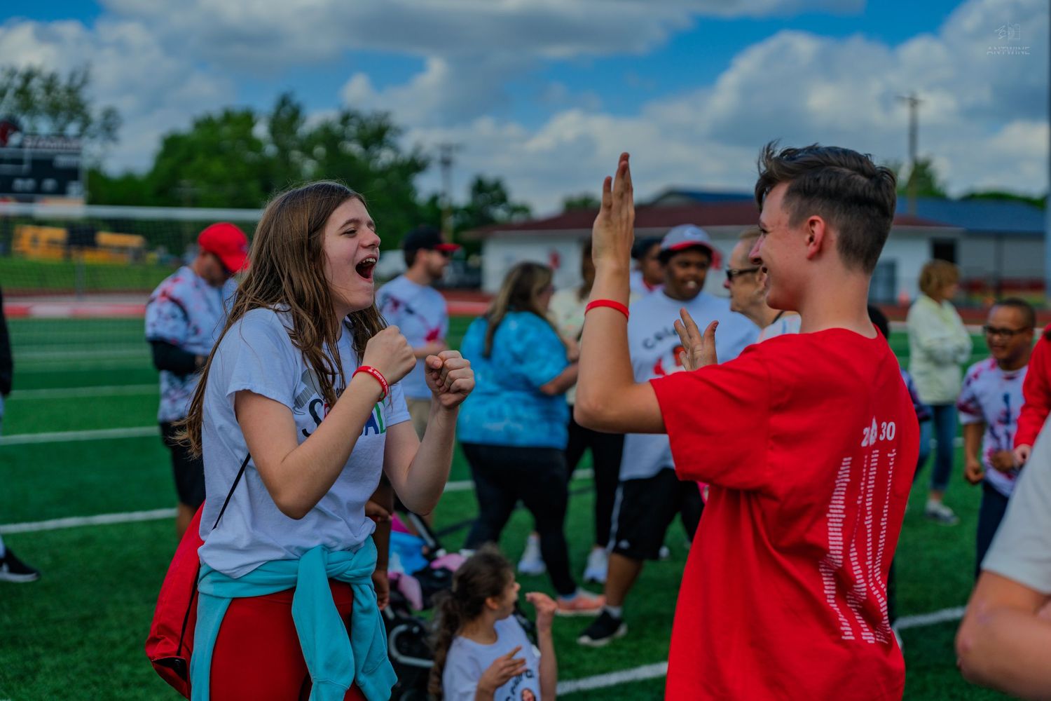Two teens high-fiving joyfully on a sports field during an outdoor community event on a cloudy day.