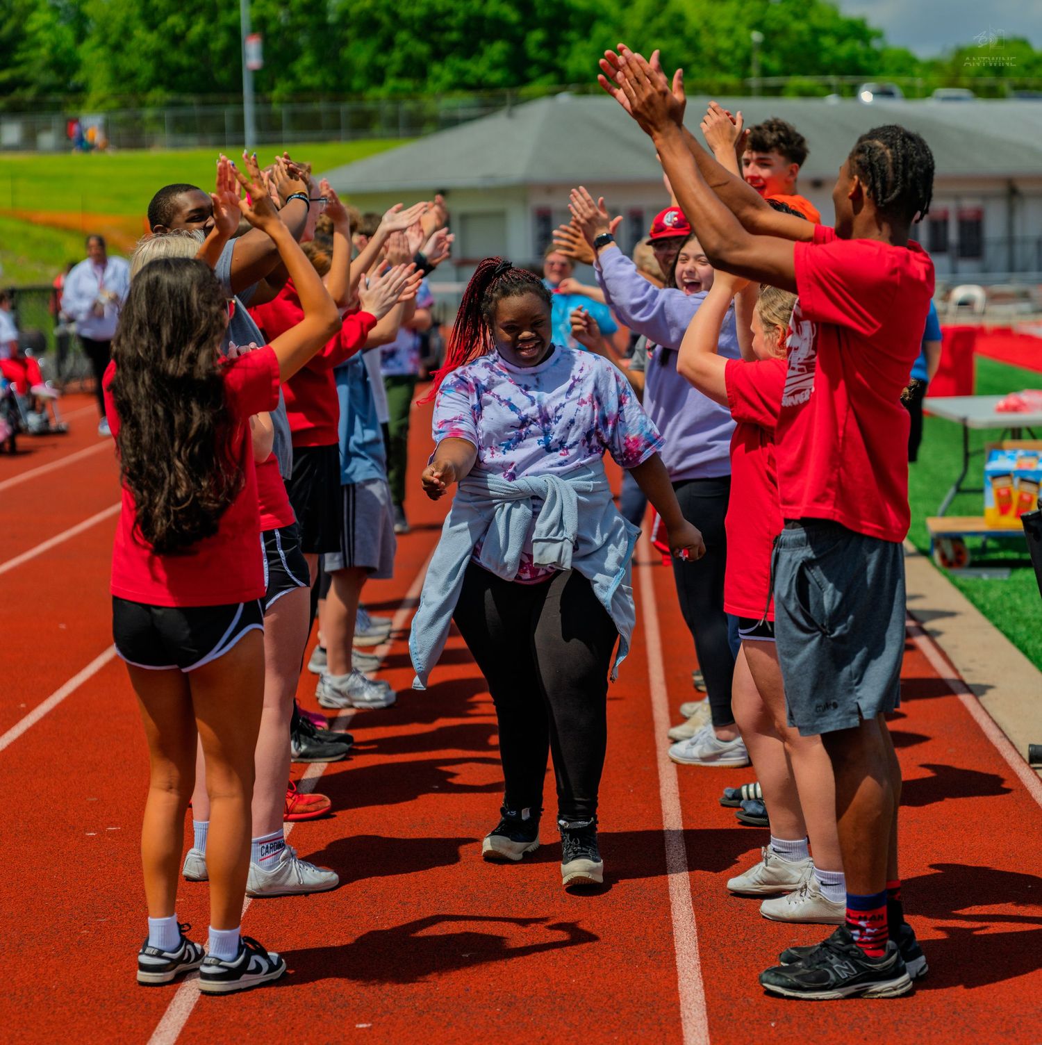 Students in red uniforms form a high-five tunnel on a track field during an outdoor sports event on a sunny day.