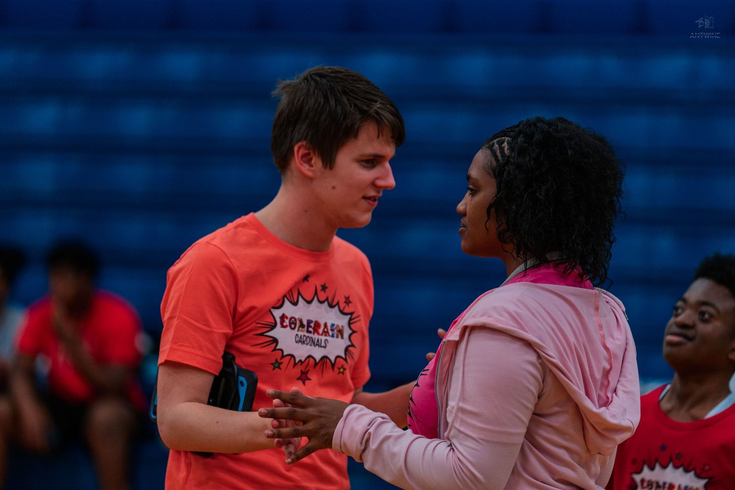 Two young people in pink and red shirts conversing at an indoor sports event.