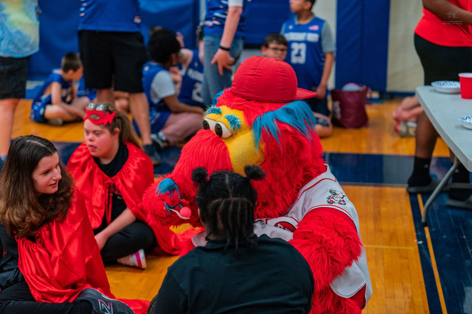 A colorful parrot mascot interacts with children in red shirts at an indoor gymnasium event.