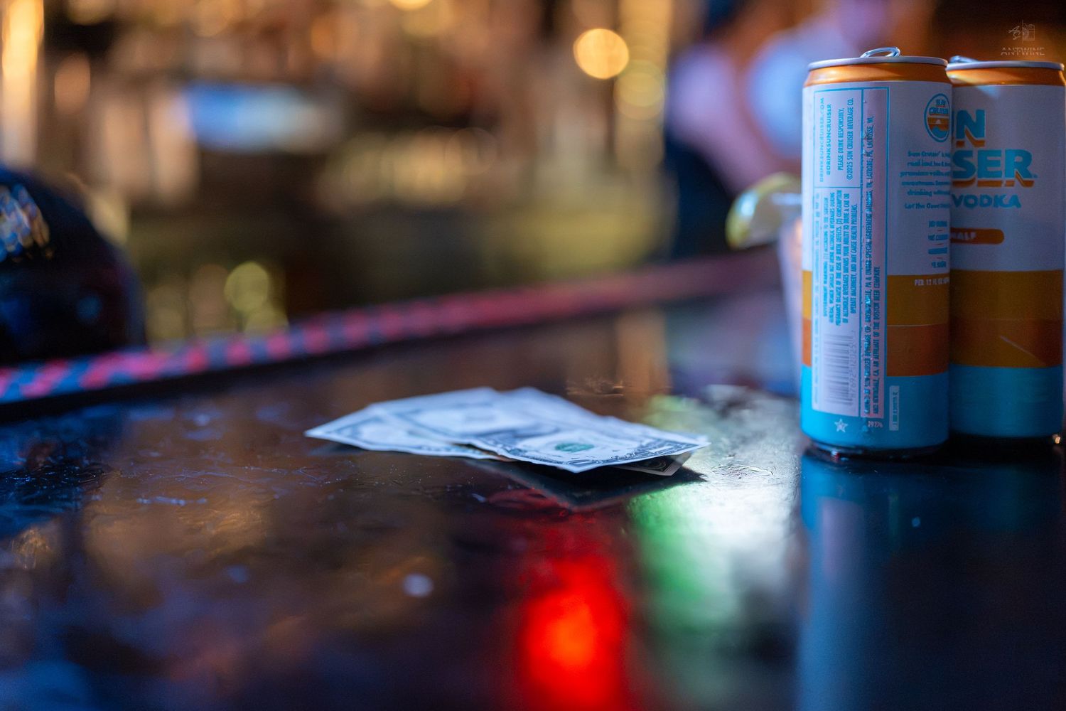 Beer cans and cash on a wet bar counter in a dimly lit bar with colorful bokeh lights.