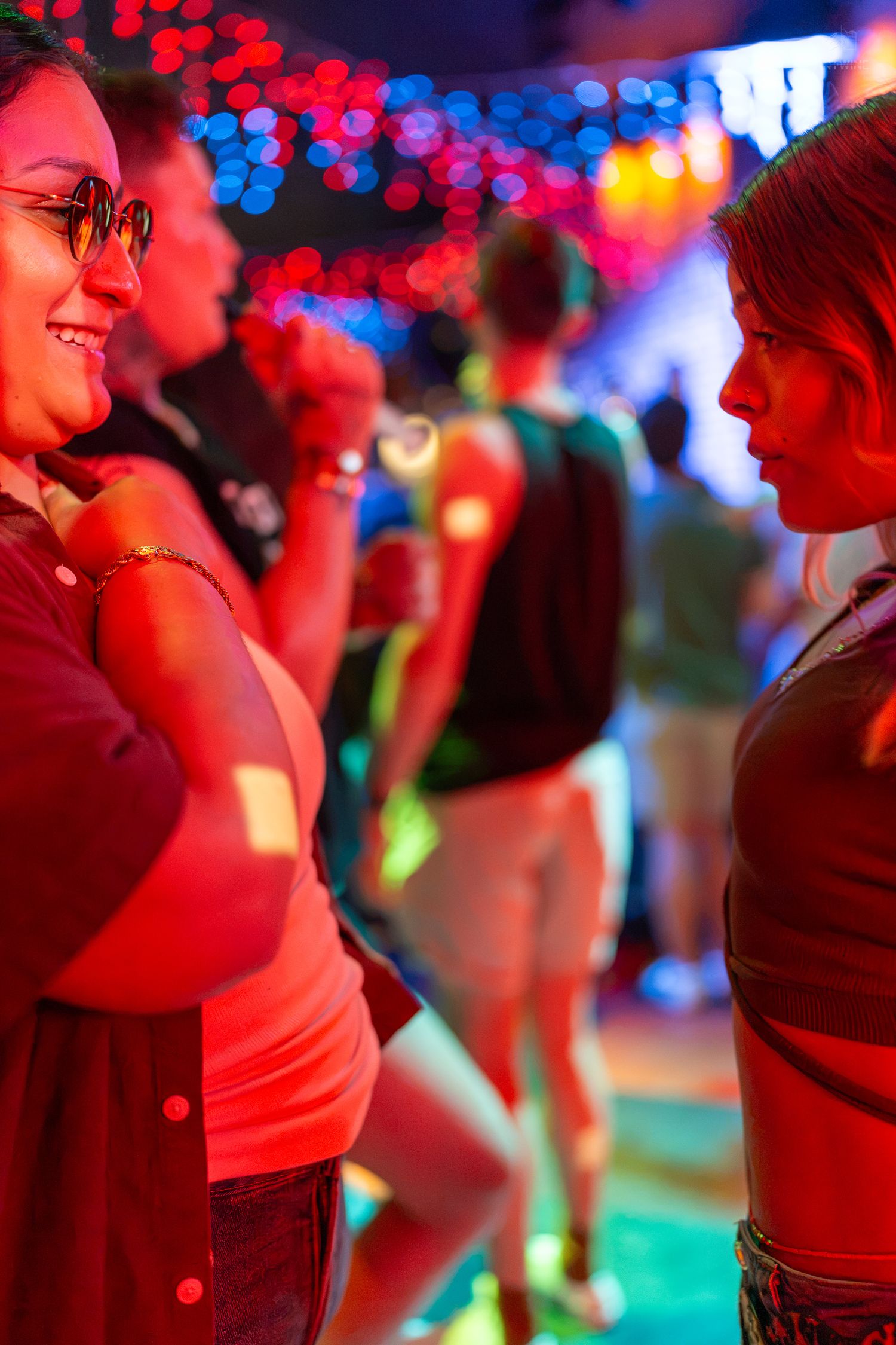 Two women conversing at a vibrant nightclub with colorful bokeh lights and red neon ambiance.