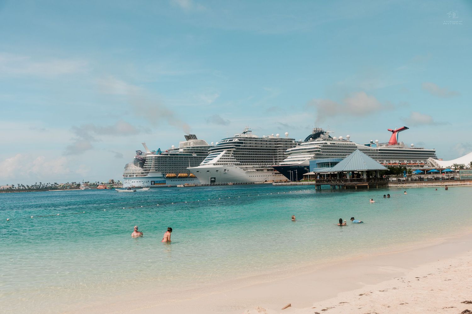Large cruise ships docked at a tropical island port with clear turquoise water and white sandy beach.