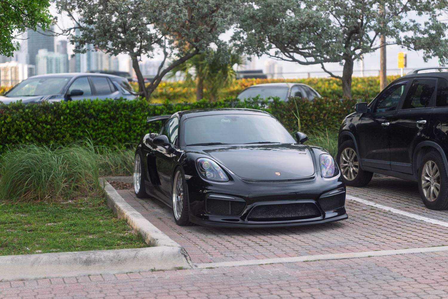 Black Porsche Cayman sports car parked on brick pavement with Miami skyline visible in background.
