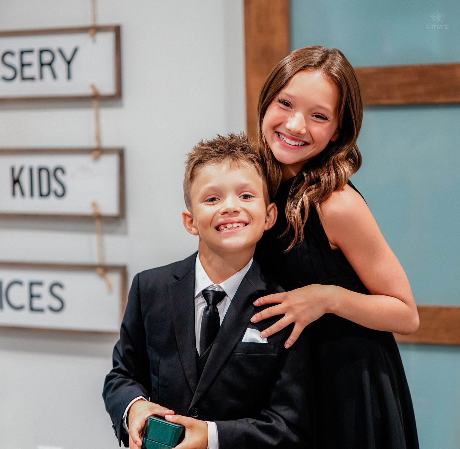 Two dressed-up children smile at a wedding, boy in black suit and girl in black dress hugging him.