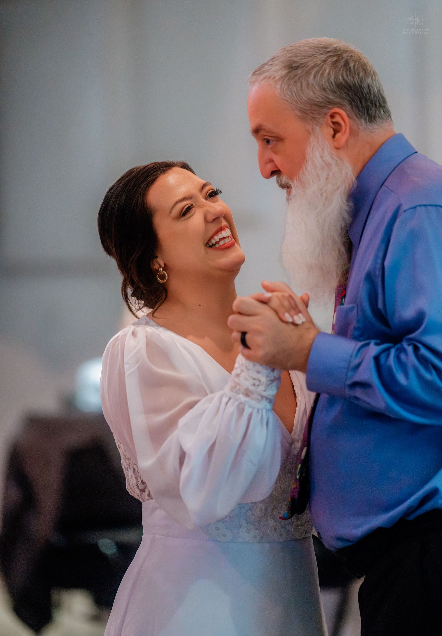 Bride in white gown laughing joyfully while dancing with bearded man in blue shirt at wedding reception.