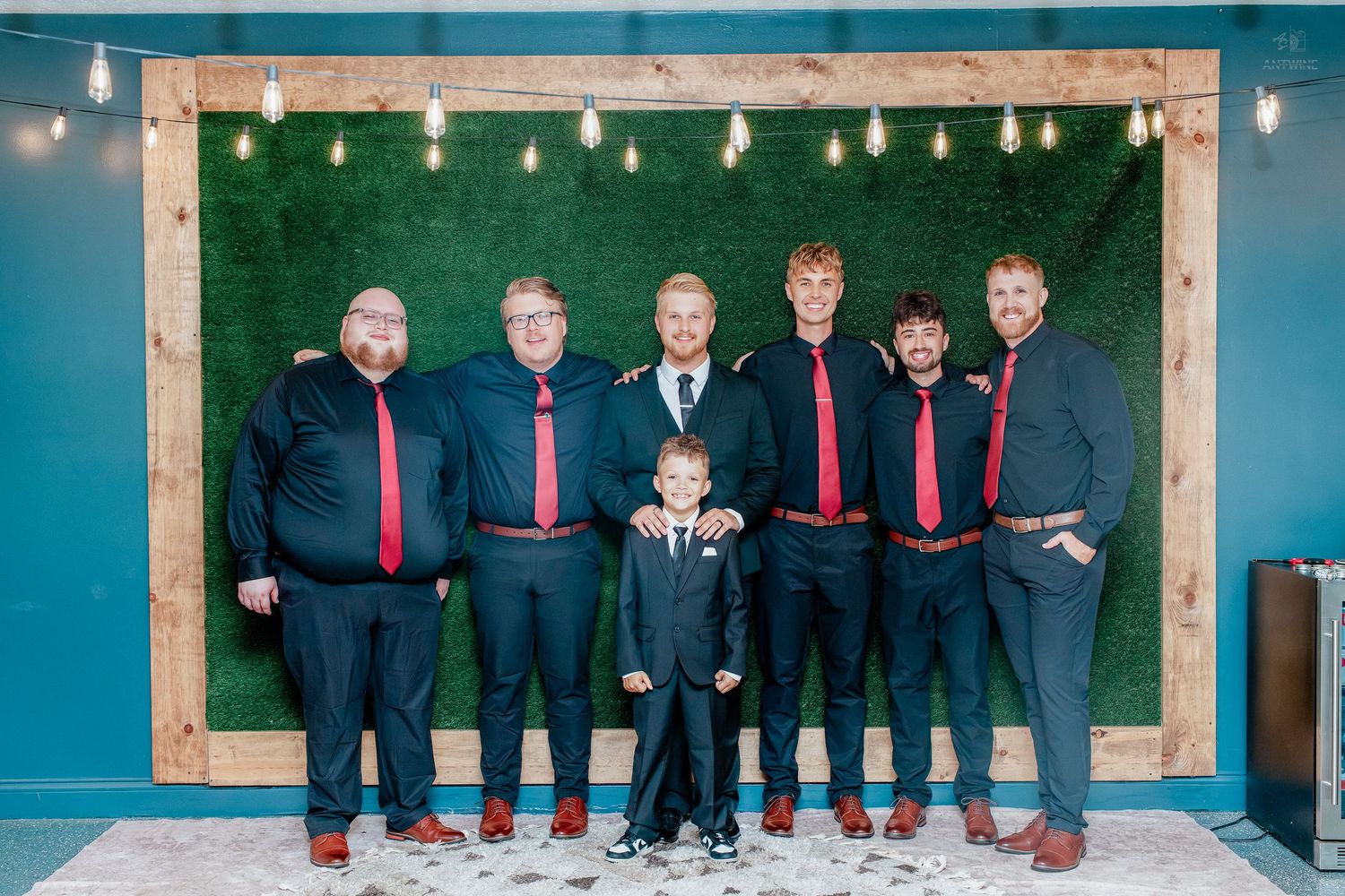 Groomsmen in black suits with red ties pose in front of a green hedge wall with Edison bulb lighting.