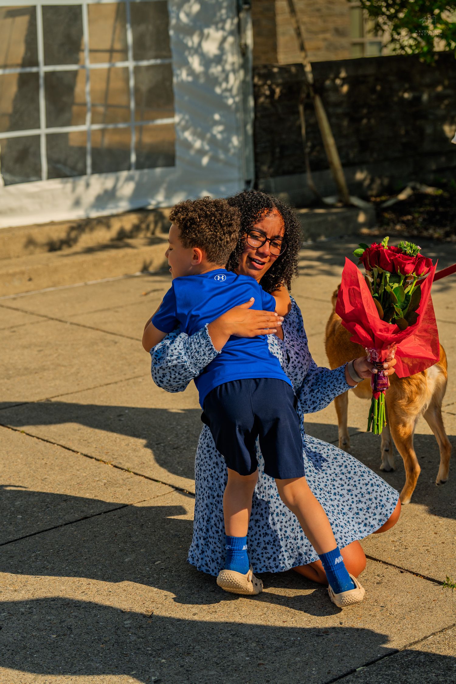 A joyful woman in a floral dress lifts a child while holding a bouquet of red roses outdoors.