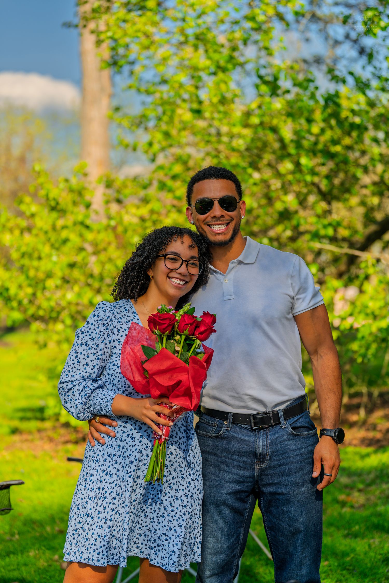 Happy couple posing outdoors with red roses bouquet in blooming garden on sunny day.