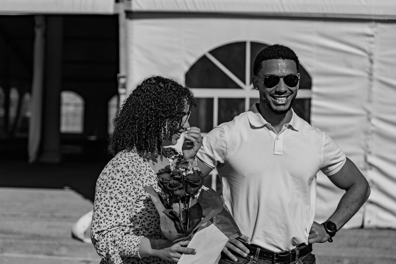 Black and white photo of a smiling man in sunglasses and a woman holding roses near a white tent.