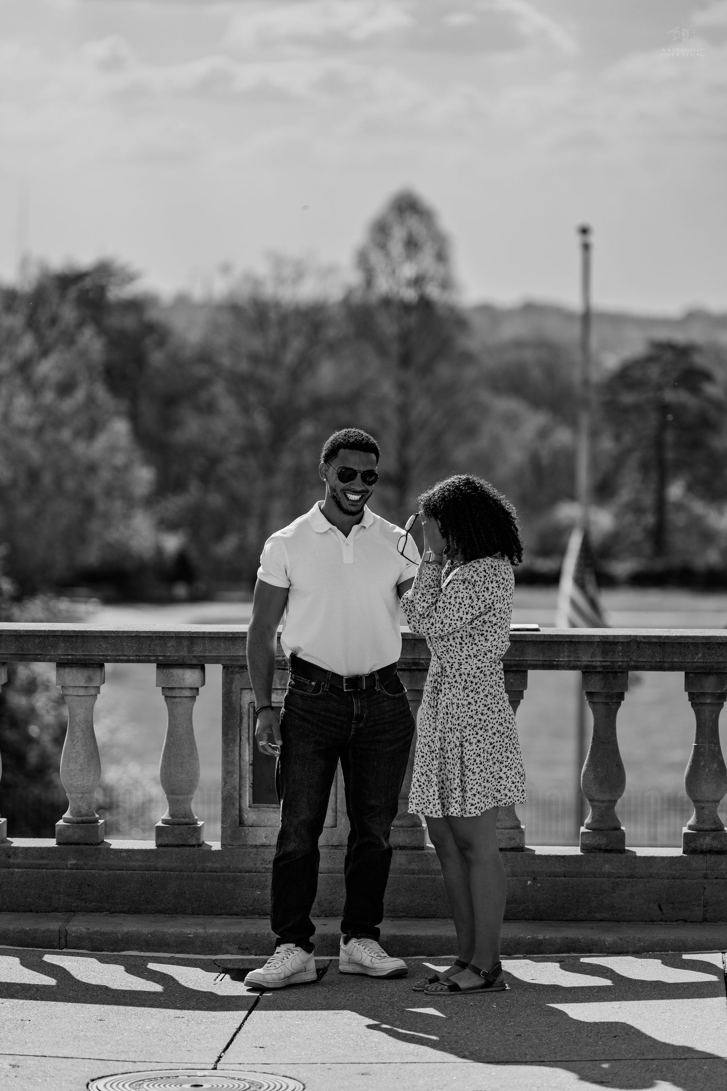Black and white photo of smiling couple standing together by stone railing in outdoor park.