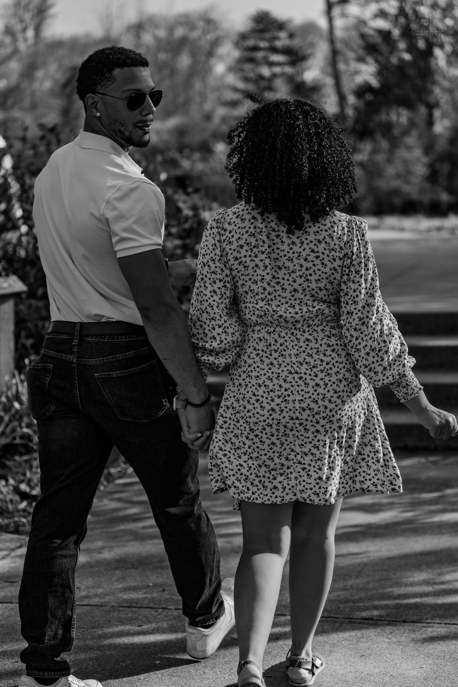 Black and white photo of a couple holding hands walking away on a path in a park.